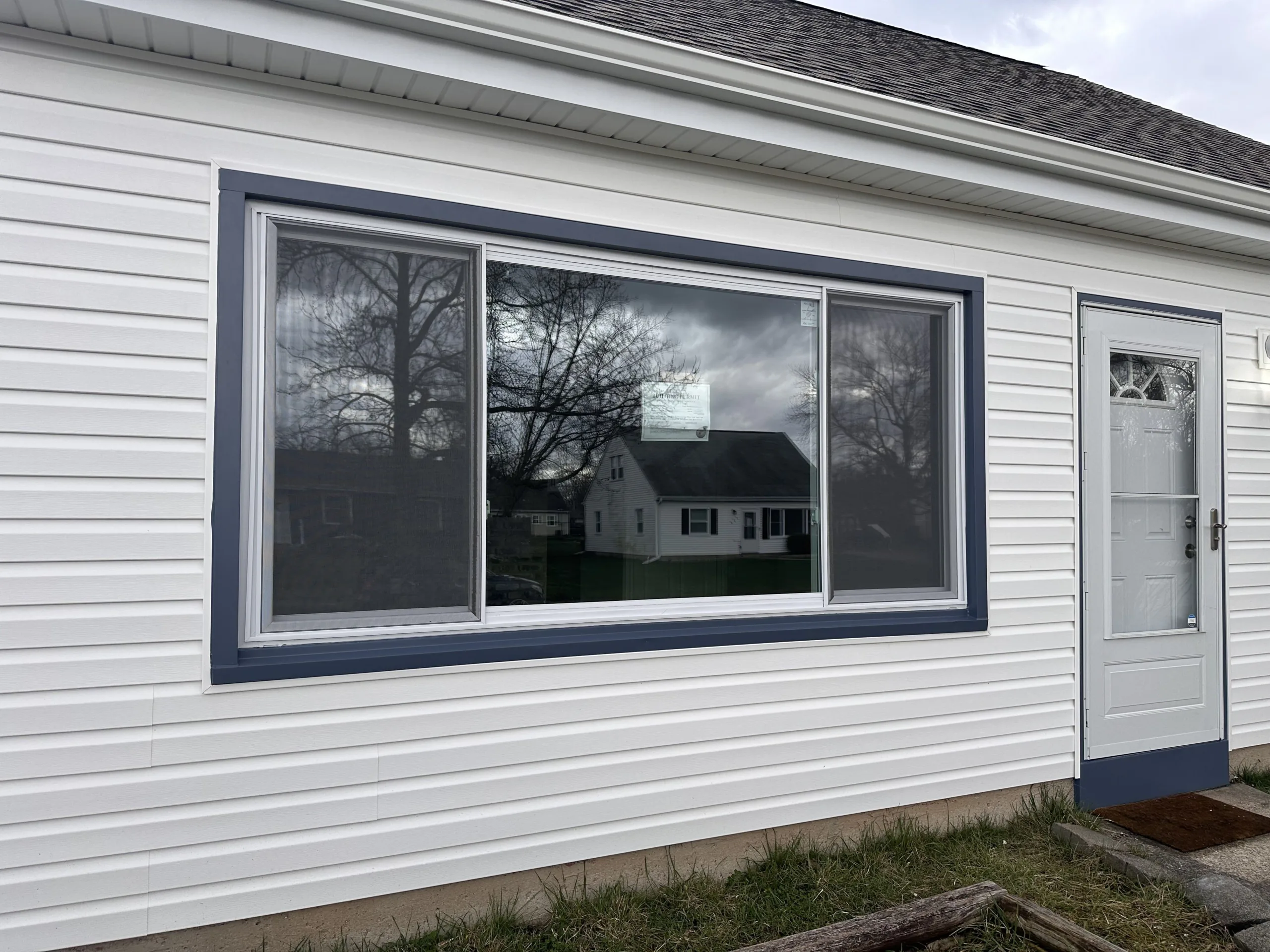 Residential facade with a large window reflecting the opposite house and a light blue entrance door.